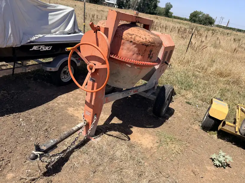 Orange towable cement mixer with its drum and hand wheel, sitting on a dirt lot.