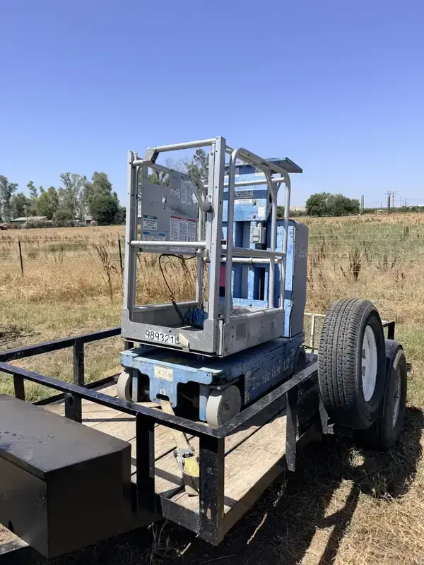 Genie GR-12 vertical mast lift loaded on a flatbed utility trailer in a grass field.