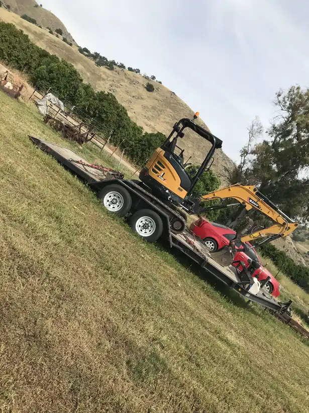 SANY mini excavator strapped to a flatbed trailer on a grass field below a dry hillside.