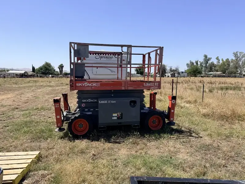 Rough-terrain scissor lift with orange guardrails standing in a dry field.